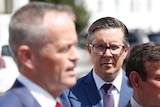 Butler wearing dark-rimmed glasses watches an out of focus Shorten speak to the media.