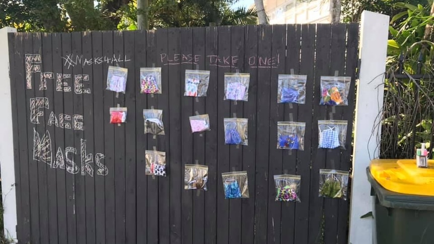 Masks packaged in zip-lock bags hang from a fence in Moorooka, Brisbane. 'Free face masks' is written in chalk on fence.