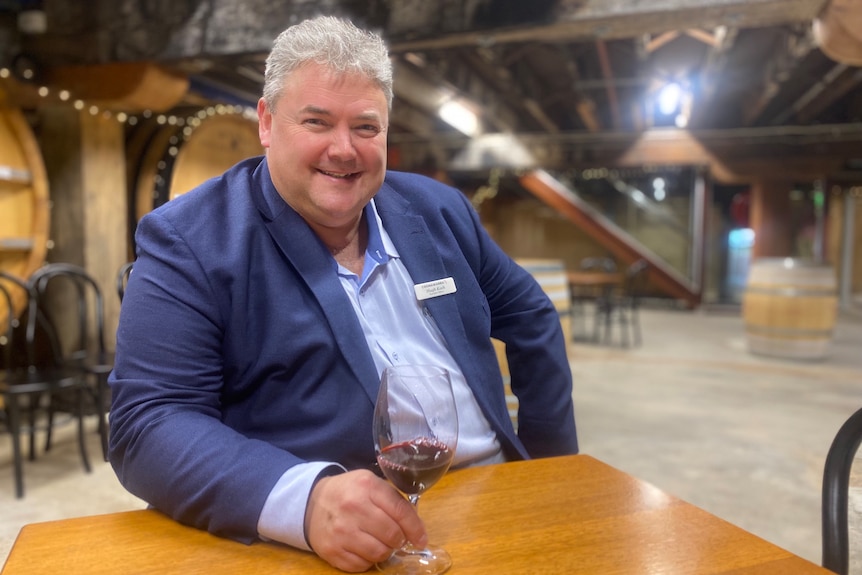 A man smiles at the camera while sitting, holding a glass of red wine. A wine cellar can be seen in the background. 