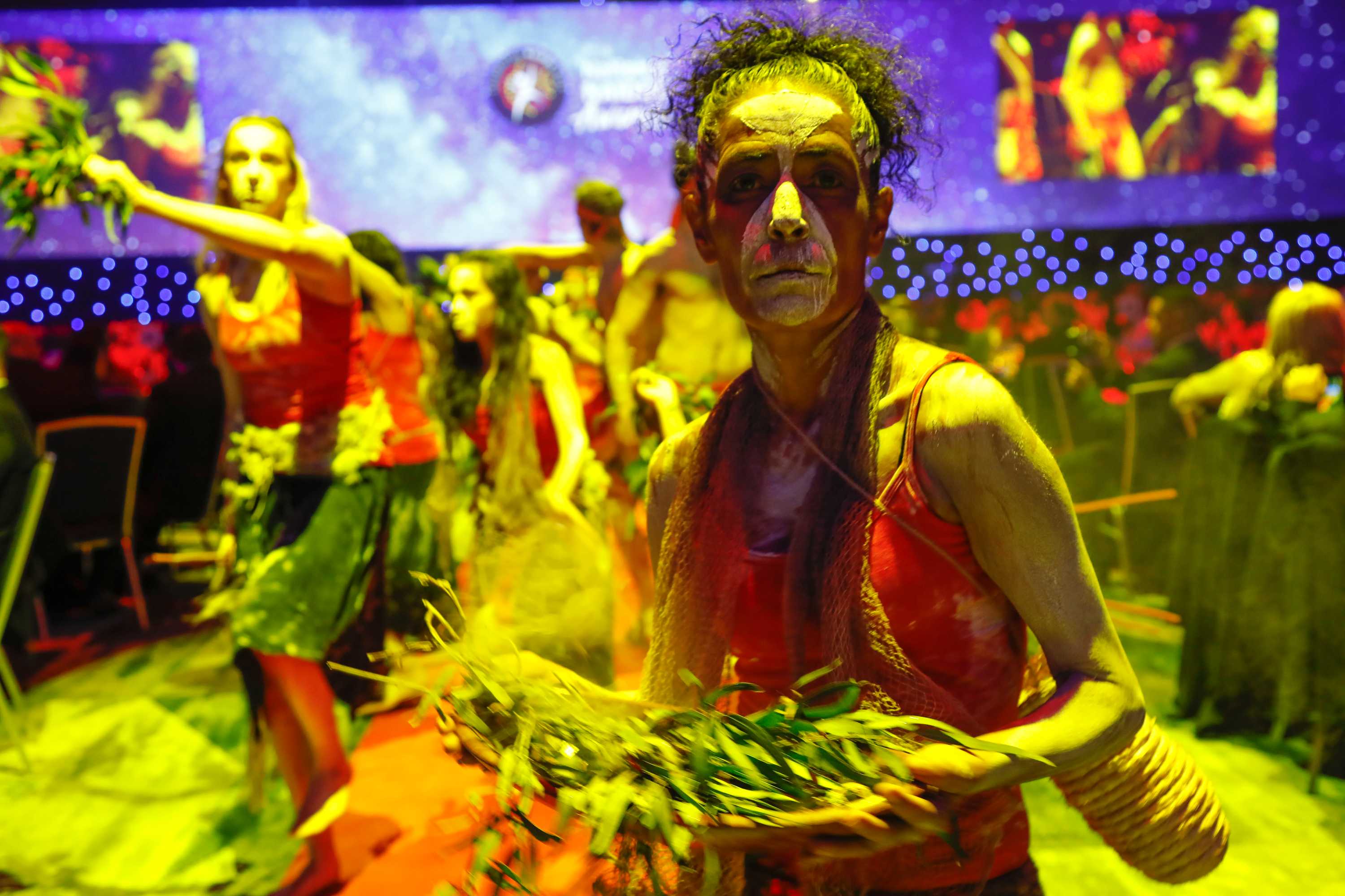 A dancer, lit with yellow light, looks into the camera, her face painted with white and eucalyptus leaves in her hand.