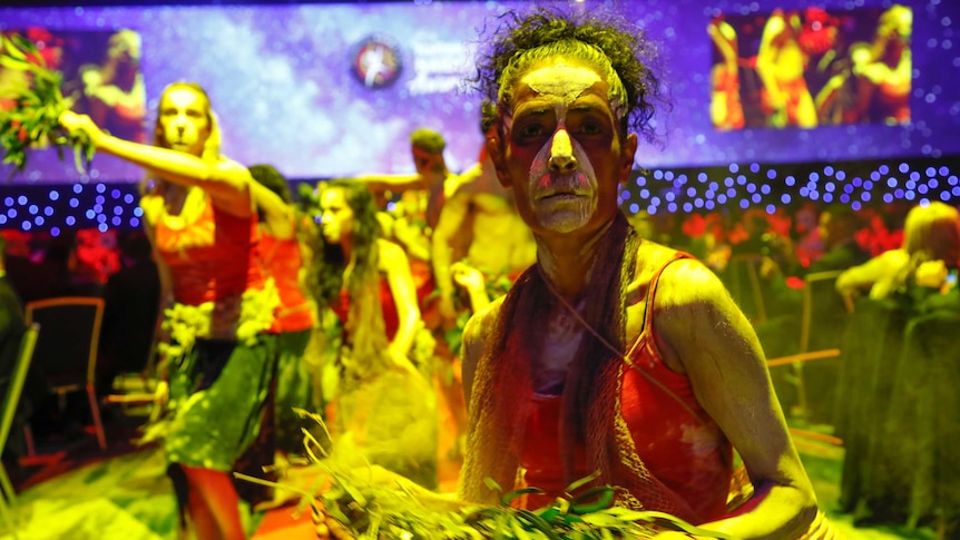 A dancer, lit with yellow light, looks into the camera, her face painted with white and eucalyptus leaves in her hand.