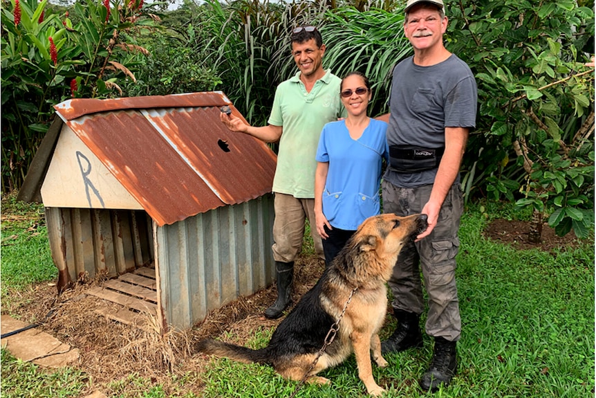 Three people stand with a German shepherd in front of a doghouse with a hole in its roof. A man is holding up a shiny stone.