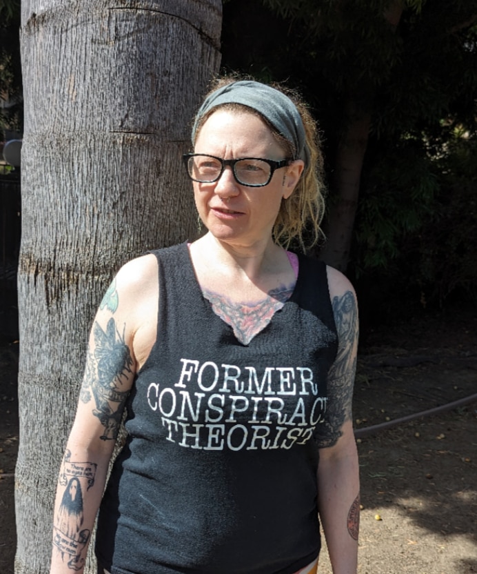 Stephanie Kemmerer, looking to side, wears singlet with words: Former conspiracy theorist. She has curly fair hair and tattooes.