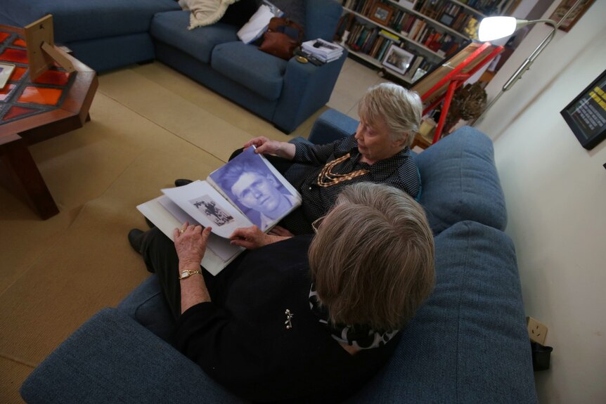 Two older women sit on a couch looking over an old portrait of their brother.