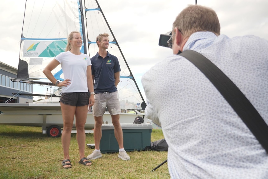 A brother and sister standing in front of a sailing boat while a photographer takes a photo