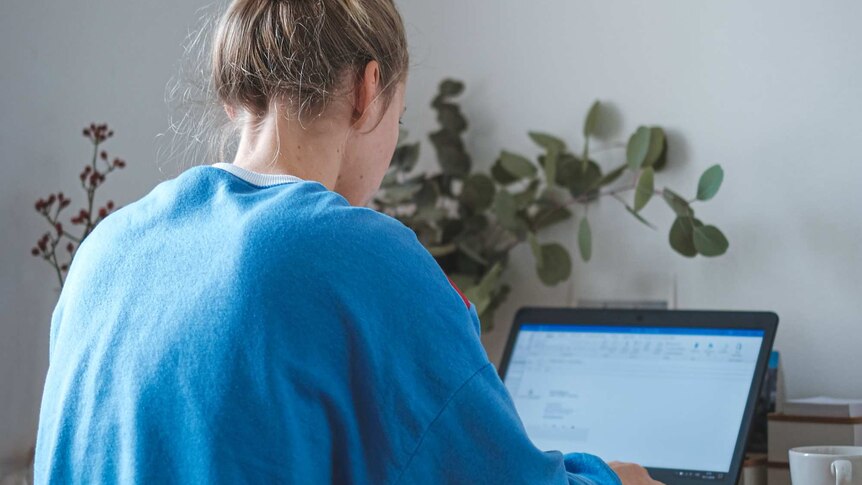 A young blonde woman in trackpants and a blue jumper sits at a desk typing on a laptop, the screen is blurred