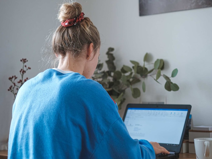 A young blonde woman in trackpants and a blue jumper sits at a desk typing on a laptop, the screen is blurred