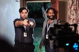 A male actor dressed as a policeman points a gun at a camera on a TV set, while another male actor looks on.