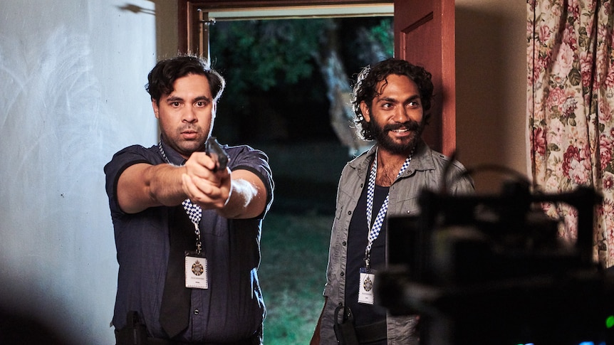 A male actor dressed as a policeman points a gun at a camera on a TV set, while another male actor looks on.