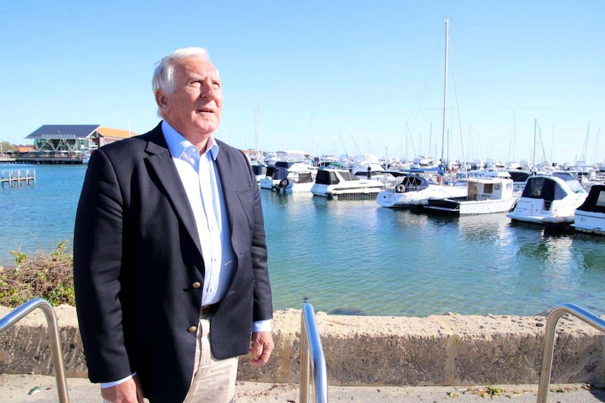 A man stands in the foreground of a marina with boats in, staring up towards a building which is out of shot.