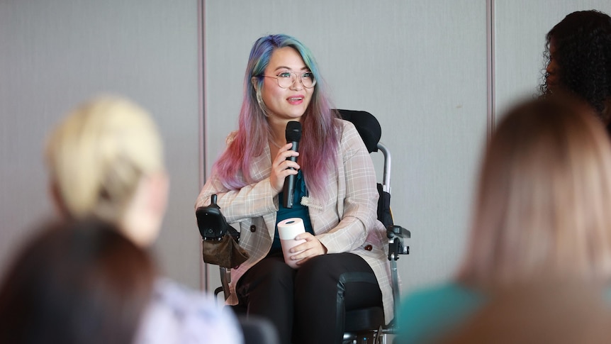 A person with long hair dyed pink and blue sits in a wheelchair with a beige jacket and black pants holding a microphone