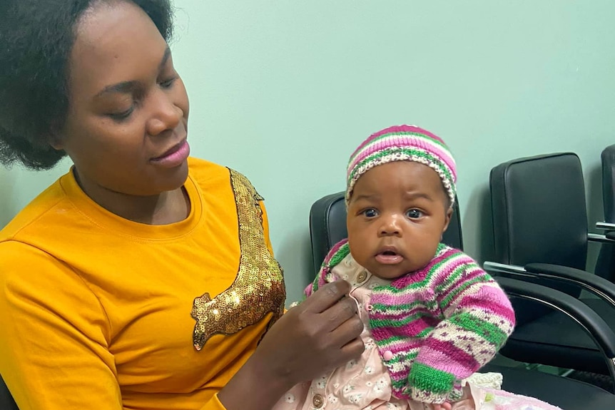 A smiling woman holds a young baby wearing colourful clothes.