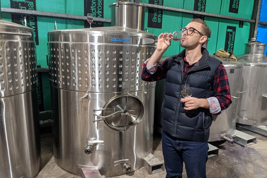 A fair-skinned man in a red chequered shirt and navy puffer vest, Mr Radny, takes a sip of red wine from a stainless steel tank.