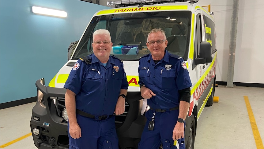 two older men in paramedic uniforms standing in from of an ambulance
