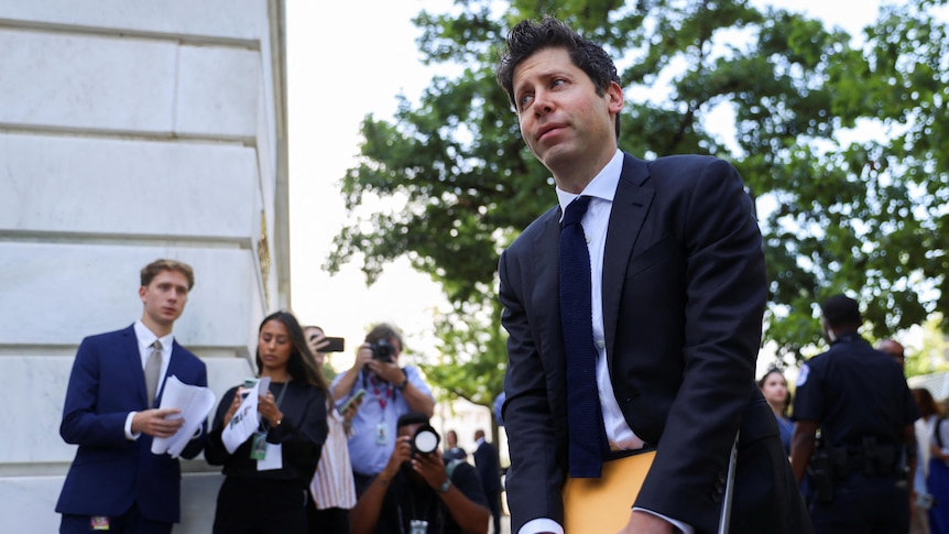 A man in a suit holding a laptop walks past a group of photographers outside 