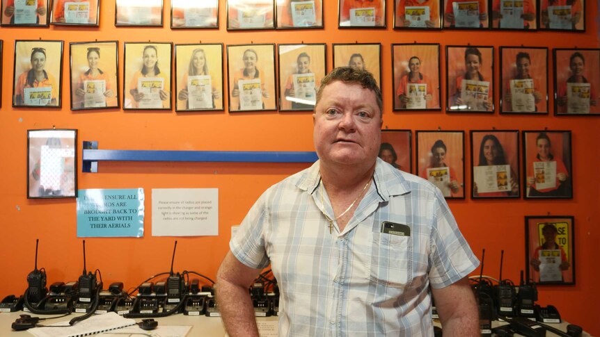 Older gentleman standing in front of bright orange wall lined with photos of traffic controllers, mostly women.