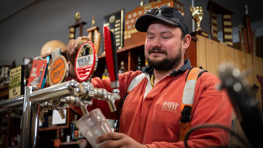 A smiling young man with a beard dressed in high vis pulls a beer from a tap from behind a bar.