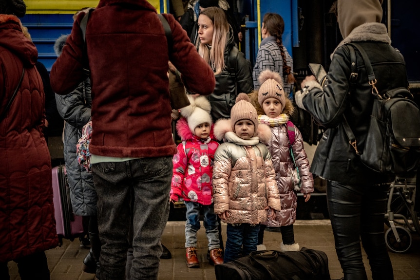 Three little girls with shiny, pink puffer jackets on a train platform. 