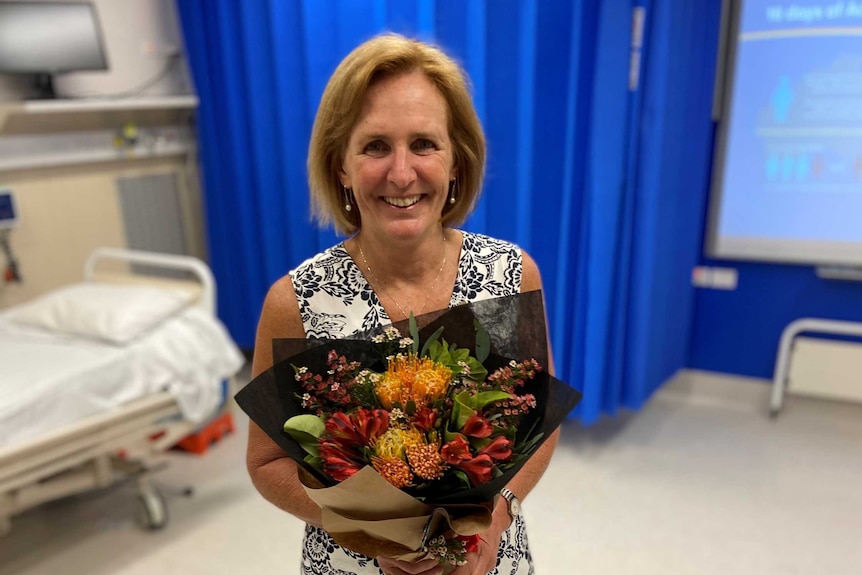 A woman stands in a hospital room wearing a dress and holding a bunch of flowers.