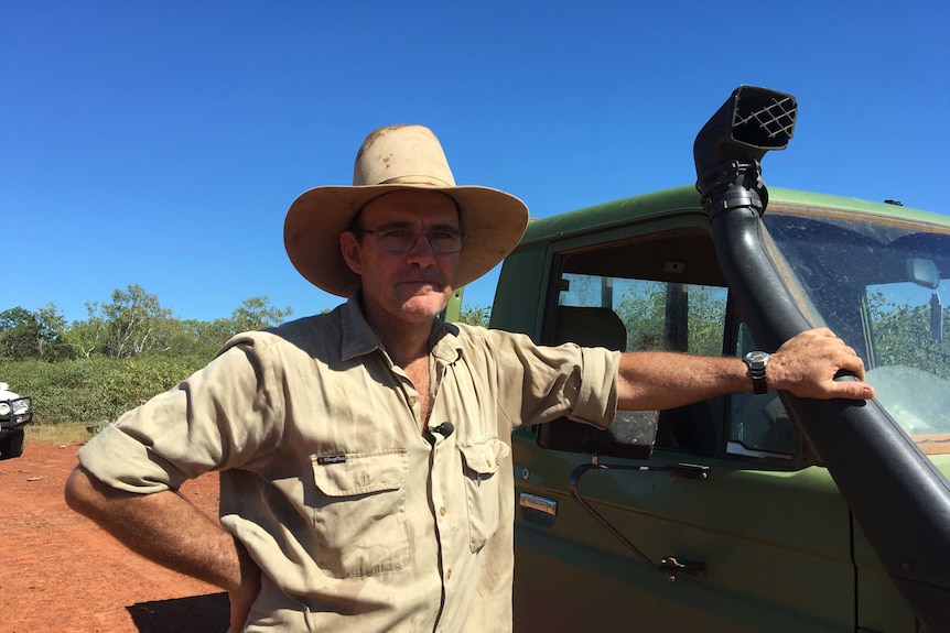 A man in a hat and work shirt leans against a ute on a cattle station.