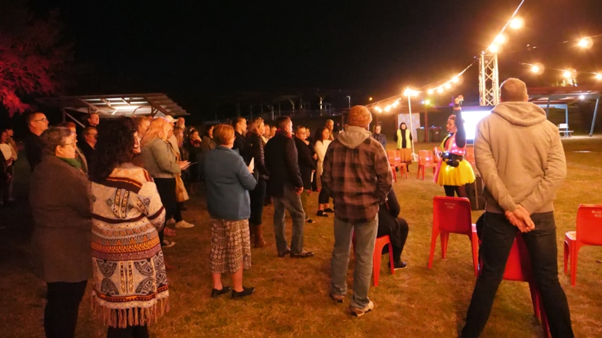 dozens of people stand in a grassy picnic area at night time, with lights strung up overhead