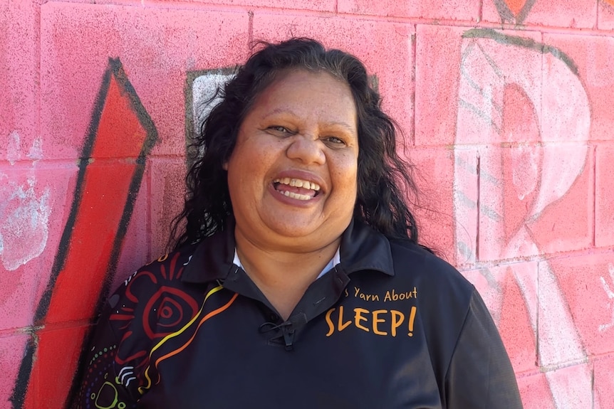 A dark skinned woman with curly hair smiles at the camera, wears tee with Indigenous art and Sleep logo.