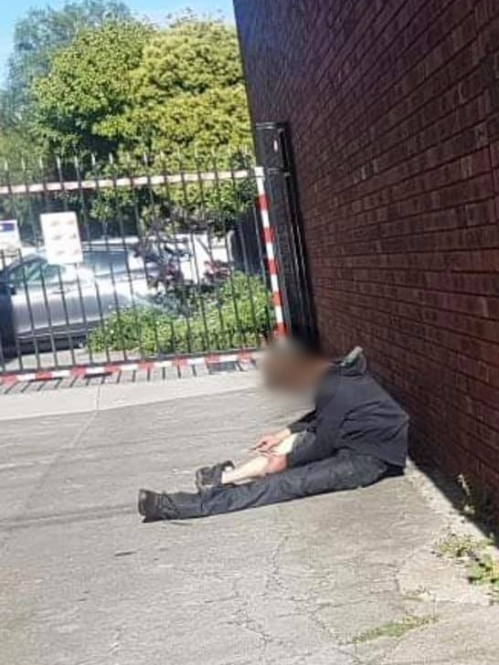 A man in black unkempt clothing slumped against a brick wall, holding what appears to be a needle near his leg.