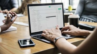 A generic image of a woman checking emails while at a table of work colleagues.