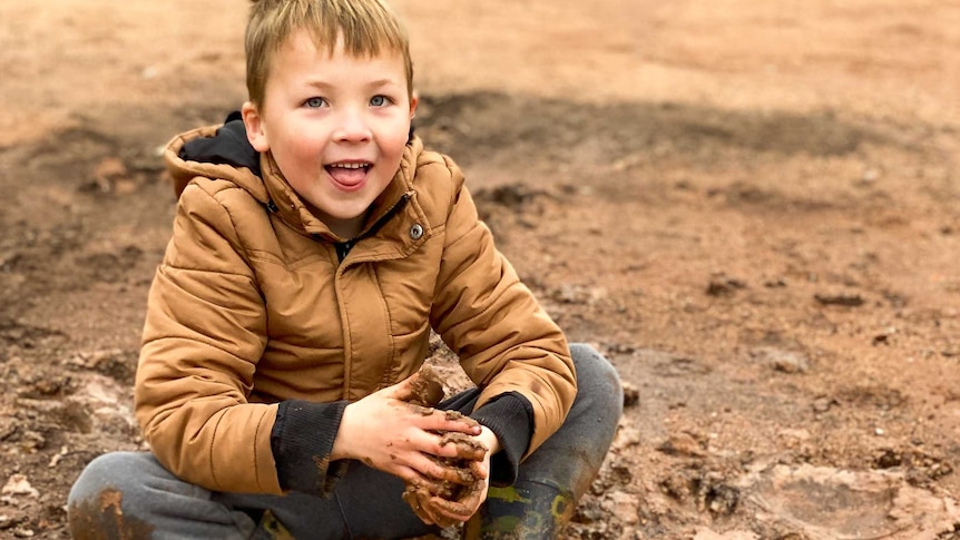 A young boy in a brown jacket and mud on his hands smiles as he sits in the dirt.