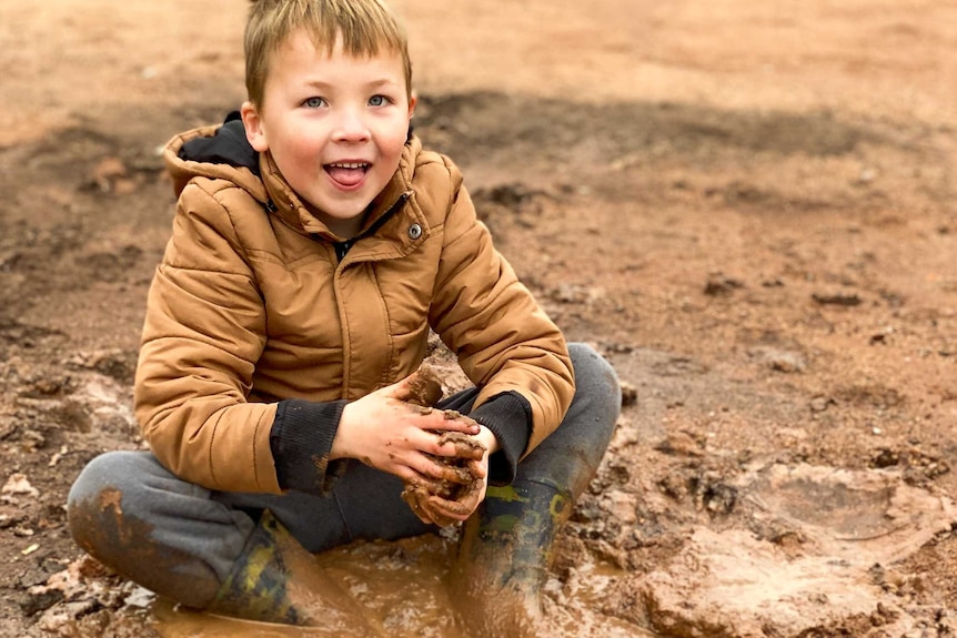 A young boy in a brown jacket and mud on his hands smiles as he sits in the dirt.