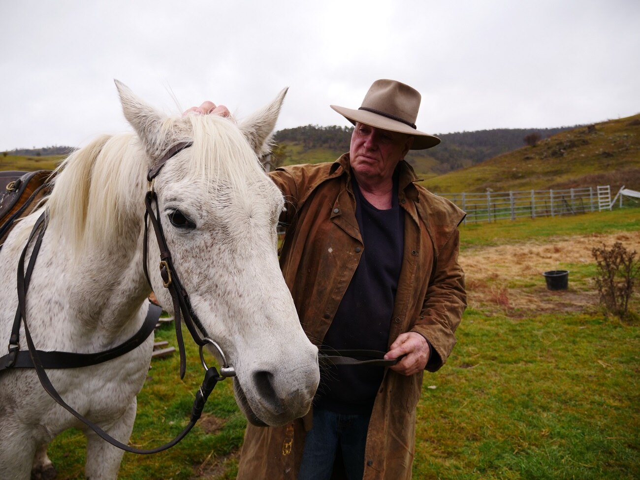 A farmer stands next to his white brumby petting its head. He wears a wide brim hat and brown oil skin jacket.