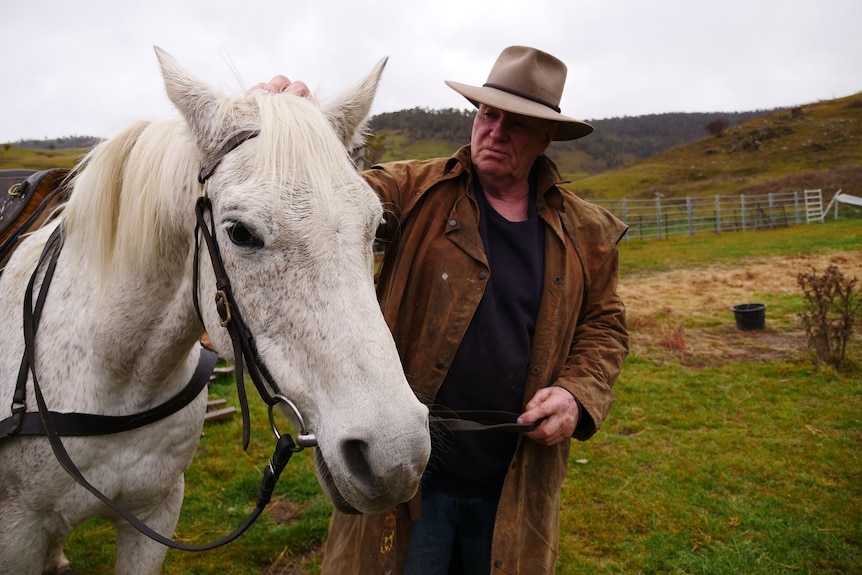 A farmer stands next to his white brumby petting its head. He wears a wide brim hat and brown oil skin jacket.