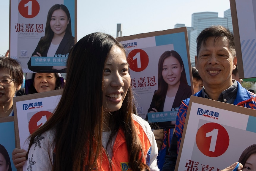 A woman wearing a high-vis orange vest smiles among a crowd of people holding signs with her face. She holds a flag