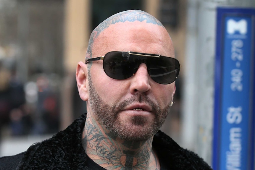 A man with tattoos on his neck and head, wearing a black coat, top and dark sunglasses.