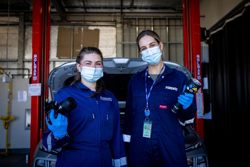 Two women wearing surgical masks, gloves and forensic overalls smile at the camera.