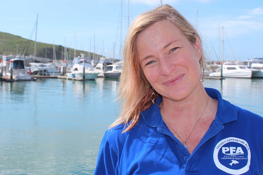 A woman, wearing a blue t-shirt, stands at a marina with boats in the background