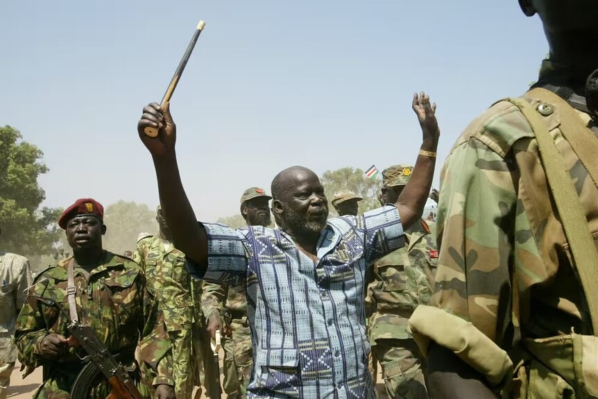 Black man in a blue striped shirt holds his hands in the air with men dressed as soldiers around him