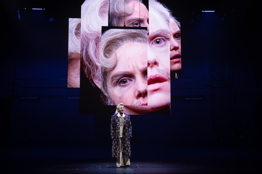 White woman dressed as man with quiffed blonde hair and moustache wears elaborate dressing gown on stage with screens behind.