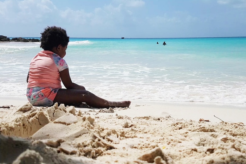 Toddler sits on white sandy Barbados beach.