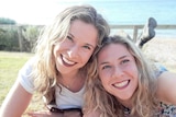 Selfie of two identical women prostrate on a grassy clifftop blurred background smiling at camera