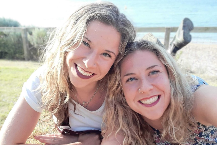Selfie of two identical women prostrate on a grassy clifftop blurred background smiling at camera