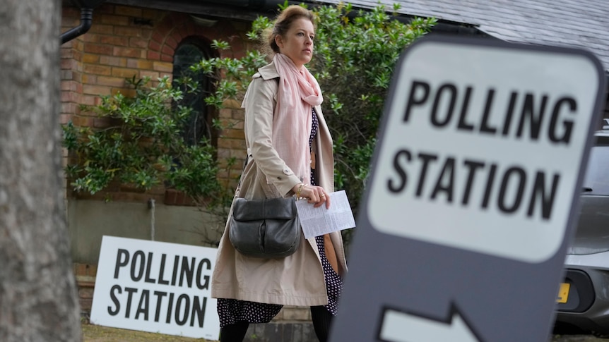 A woman holds a document as walks on pavement past signs saying "polling station".
