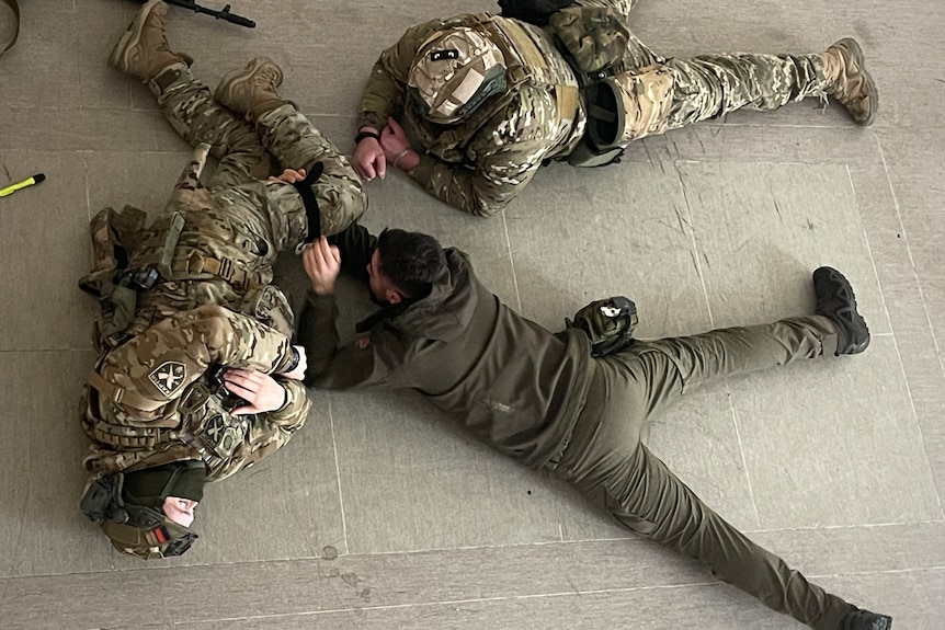 Men in army fatigues lie on the floor of a room during a training exercise.