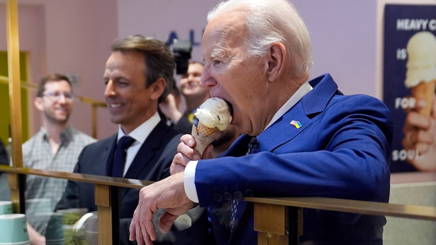 Joe Biden leans over a counter eating a vanilla icecream while surrounded by aides.