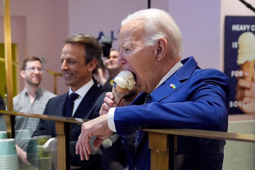 Joe Biden leans over a counter eating a vanilla icecream while surrounded by aides.