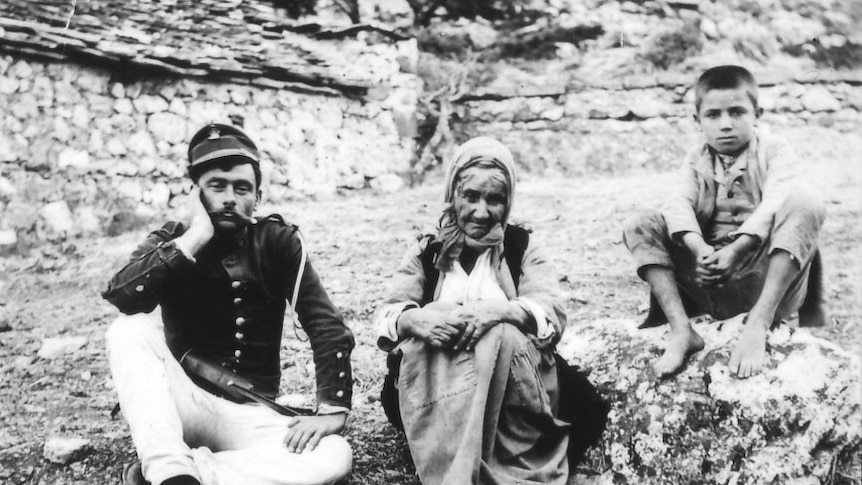 Three people sit on the ground: a mustachioed man in uniform, an elderly woman and a boy.