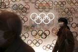 A women wearing a face mask stands in front of a series of multiple Olympic rings.