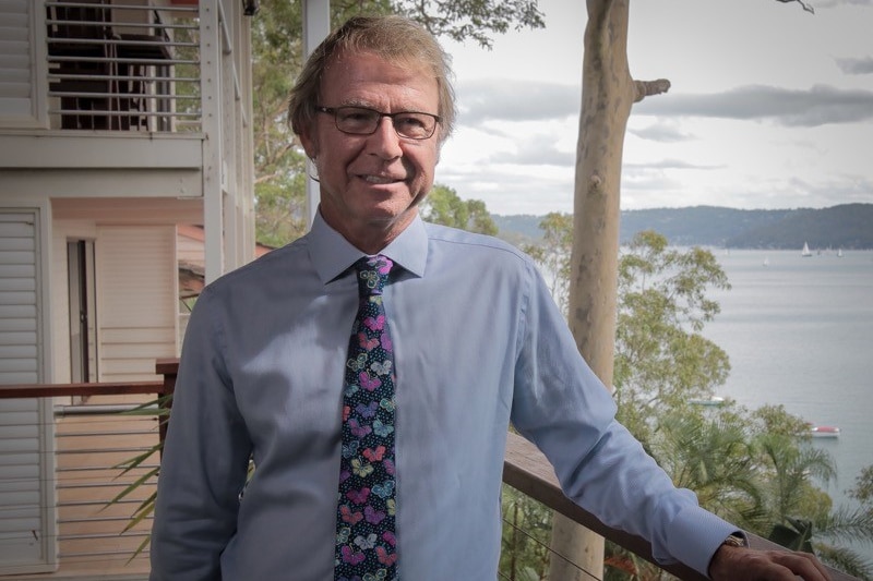 A man wearing a light blue shirt and tie with colourful butterflies on it stands on a verandah overlooking water.