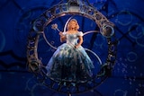 A Black woman in an ornate blue gown, including a wand, grins brightly, as she stands on stage in a circular set piece