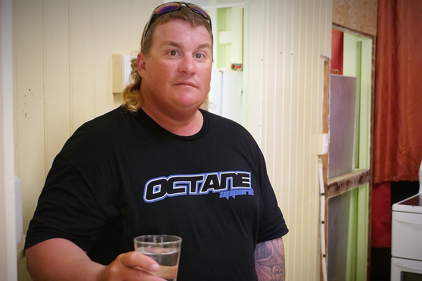 A man standing in the kitchen holding a glass of water looking at the camera